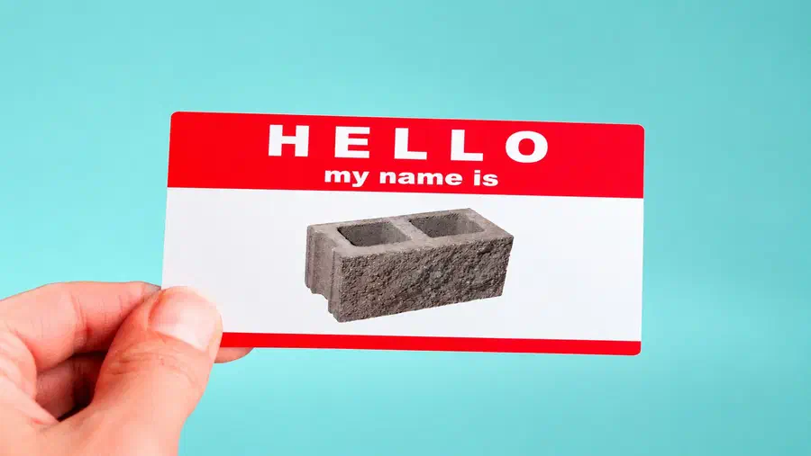 A hand holding a red-and-white name tag that reads “Hello my name is,” featuring a concrete cinder block in place of a person's name against a light blue background
