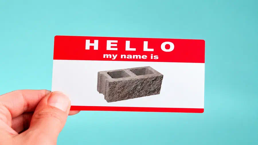 A hand holding a red-and-white name tag that reads “Hello my name is,” featuring a concrete cinder block in place of a person's name against a light blue background