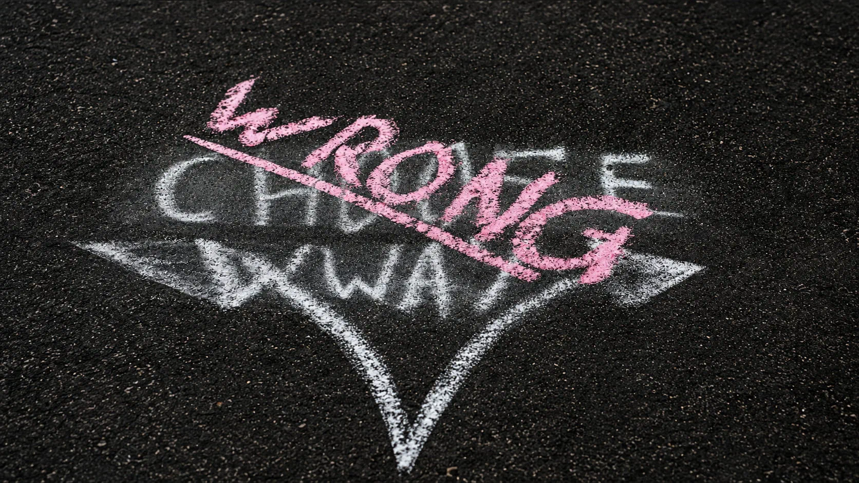 Chalk graffiti on dark pavement showing the words “Wrong Way” crossed over a large white directional arrow